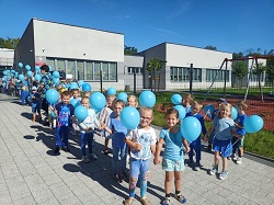 Dzieci ubrane na niebiesko idą przed przedszkolem. W ręku trzymają niebieski balon