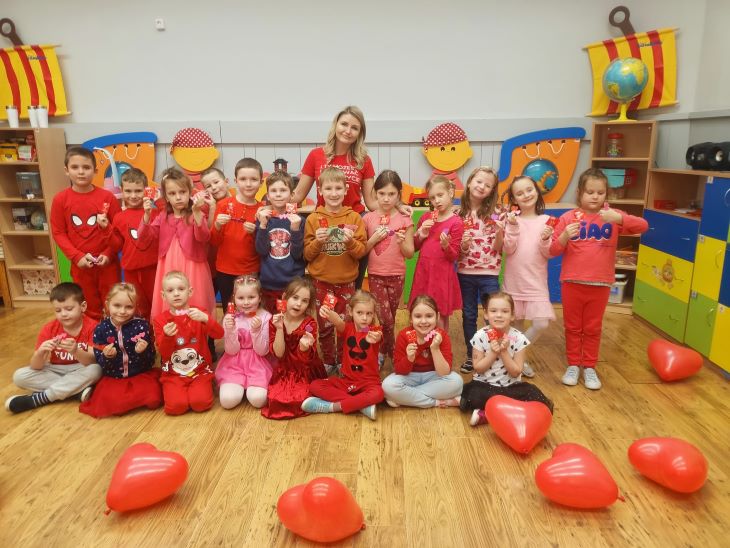 Dzieci ubrane na czerwono stoją w sali. Na ziemi leżą balony w kształcie serc. 