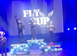Na zdjęciu widać młodą dziewczynę stojącą na scenie podczas uroczystej gali lub zawodów. Trzyma wysoko uniesiony puchar oraz dyplom, co świadczy o jej zwycięstwie. Za nią znajduje się duże, podświetlone logo „FLY CUP”. Scena jest oświetlona kolorowymi światłami, a po obu stronach umieszczone są dekoracje z niebieskich balonów. Dziewczyna wygląda na dumną i szczęśliwą z osiągniętego sukcesu.