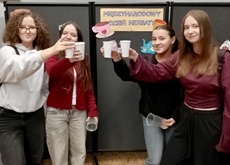 Na zdjęciu widać cztery uczennice stojące obok siebie, które wznoszą toast papierowymi kubeczkami z herbatą. Dziewczęta są uśmiechnięte, a cała scena oddaje miłą i radosną atmosferę wspólnego świętowania. W tle znajduje się gazetka tematyczna z napisem „Międzynarodowy Dzień Herbaty” oraz dekoracje nawiązujące do tego wydarzenia, co wskazuje, że zdjęcie zostało wykonane podczas szkolnych obchodów tego święta. Całość podkreśla integrację uczniów oraz przyjemny charakter spotkania.