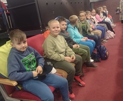 Na zdjęciu widać grupę dzieci siedzących w rzędzie na sali, prawdopodobnie przed rozpoczęciem przedstawienia teatralnego. Uczniowie są uśmiechnięci i spokojnie czekają, trzymając swoje plecaki. Atmosfera wygląda na radosną i pełną oczekiwania.