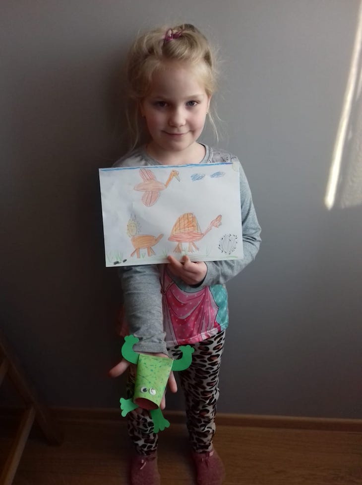 dziewczynka prezentuje obrazek z dinozaurami oraz zrobioną z rolki po papierze toaletowym zieloną żabkę