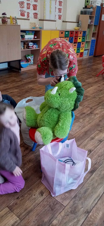 na zdjeciu pani pediatra bada dużego, zielonego, pluszowego dinozaura