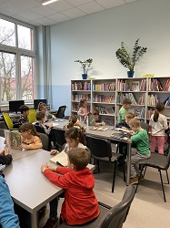 na zdjęciu siedzą w bibliotece przy dwóch długich stołach, w tle widać regały pełne książek, dzieci przy stołach przeglądają książki