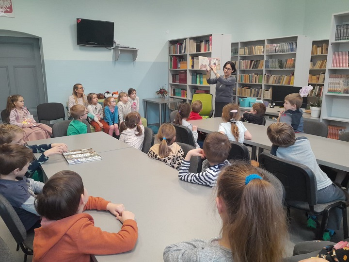 na zdjęciu dzieci w bibliotece siedzą przy dwóch długich stołach, na drugim planie bibliotekarka czyta dzieciom opowiadanie