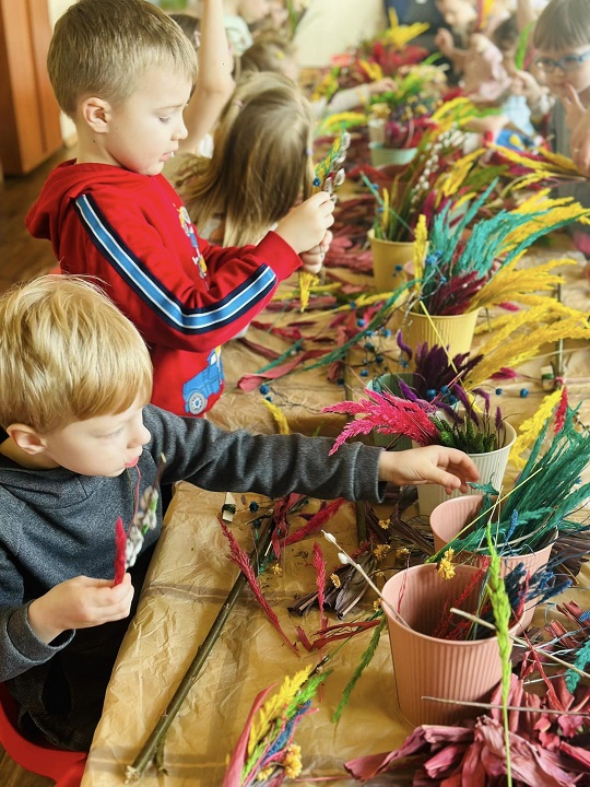 na zdjęciu dzieci siedzą przy długim stole na którym leżą materiały dekoracyjne do zrobienia palm wielkanocnych, gałązki, bazie, wstążki, kolorowe trawy
