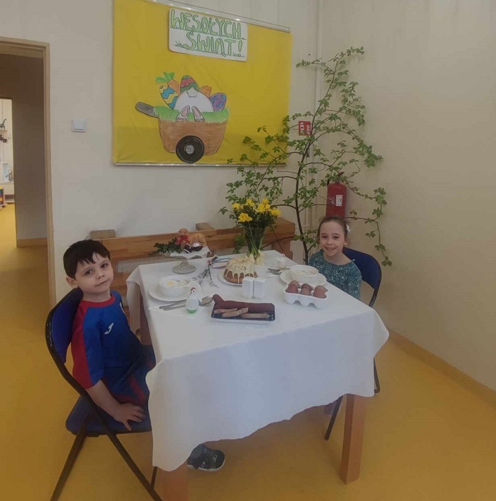 na zdjęciu dwoje dzieci siedzi przy stole z wielkanocną dekoracją , jajka, wędlina , kwiaty, babka i mazurek