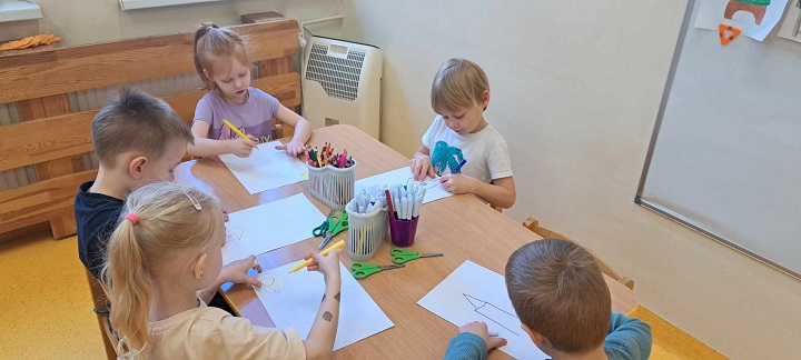 Na zdjęciu piątka dzieci siedzi przy stoliku. Rysują na kartce papieru domy.
