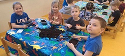 Na zdjęciu czwórka dzieci siedzi przy stoliku. Wkładają ziemię do plastikowych pudełeczek.