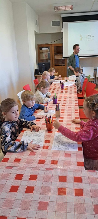 Na zdjęciu dzieci siedzą przy stole i zajmują się kolorowaniem otrzymanych obrazków. W tle znajduje się włączony rzutnik.