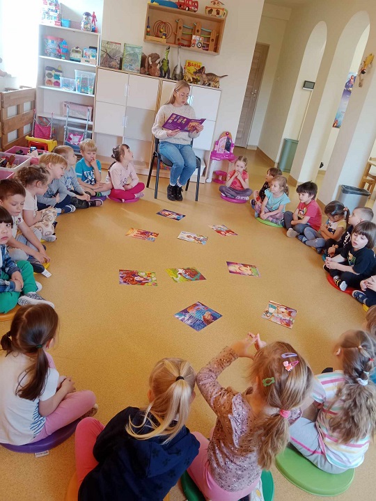 dzieci siedzą w kole na podłodze, w środku koła leżą książki, dorosła osoba czyta dzieciom bajkę