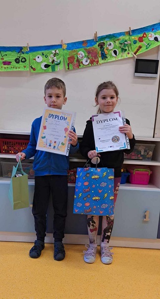 na zdjęciu stoi dwójka dzieci, chłopiec i dziewczynka, w dłoniach trzymają dyplomy oraz torebki prezentowe.