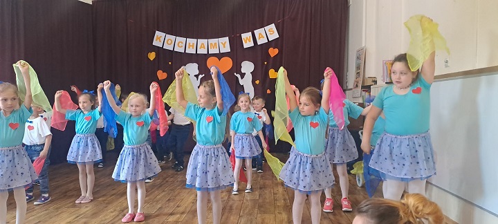 Na zdjęciu widać dzieci, które tańczą i pokazują gestem słowa piosenki. W tle znajduje się kotara z dekoracją.
