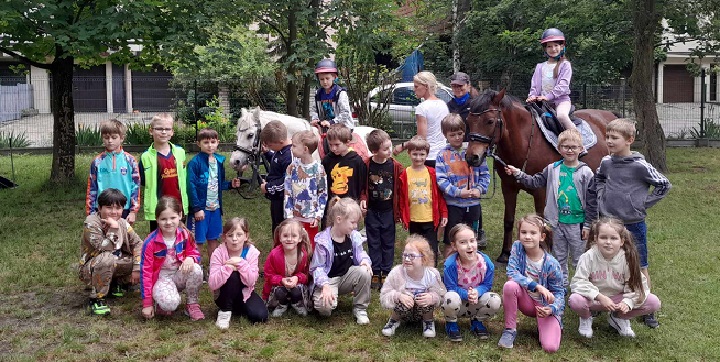 na zdjęciu grupa dzieci stoi w dwóch rzędach, w tle dwójka dzieci siedzi na koniach