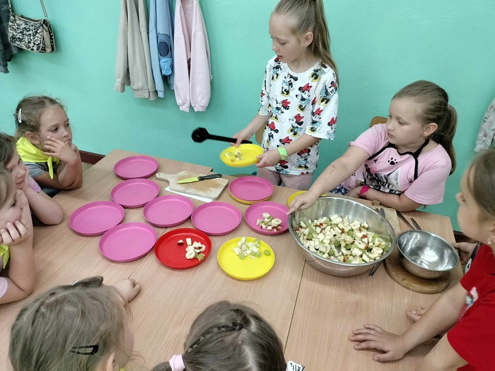 na zdjęciu dzieci przy stole robią owocową sałatkę, na stole leży dużo różowych talerzyków