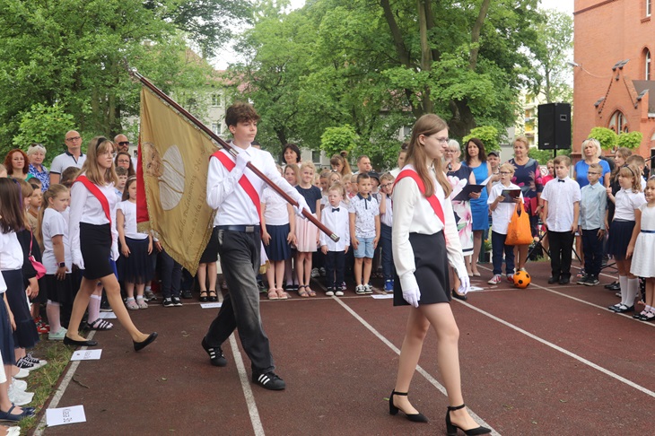 Uczniowie stpoją w czasie apelu przy okazji zakończenia roku szkolnego 2023/24 na pierwszym planie poczet sztandarowy