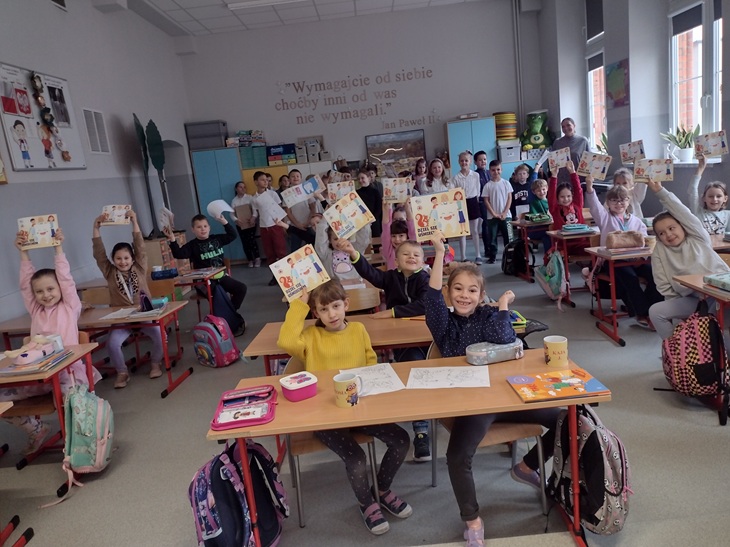 Na przesłanym zdjęciu widać grupę uczniów siedzących w klasie przy ławkach. Dzieci radośnie podnoszą do góry trzymane w rękach książeczki lub inne materiały, co wskazuje na udział w wydarzeniu lub akcji edukacyjnej, prawdopodobnie związanej z obchodami Dnia Uśmiechu. 