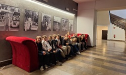 Na zdjęciu widać grupę dzieci siedzących na długiej, czerwonej kanapie ustawionej pod ścianą. Kanapa ma nietypowy, zaokrąglony kształt po bokach. Dzieci są ubrane elegancko — wiele z nich ma koszule, swetry lub marynarki. Wydają się być w podobnym wieku, prawdopodobnie uczniowie szkoły podstawowej.