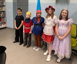 Na zdjęciu widać grupę pięciorga dzieci stojących w szeregu, ubranych w kolorowe stroje bajkowych postaci 🎭. Znajdują się w sali szkolnej, prawdopodobnie w bibliotece – w tle widać regał z książkami
