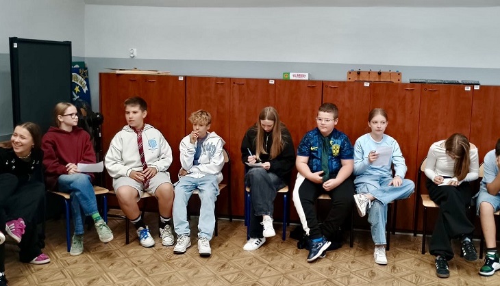 Na zdjęciu widzimy grupę młodzieży siedzącą w półokręgu na krzesłach w klasie szkolnej. W tle znajdują się drewniane szafki oraz tabliczka z napisem „SŁUŻBOWE”. Większość osób ma w rękach kartki lub zeszyty i coś zapisuje. Panuje luźna atmosfera – część osób się uśmiecha, inni są skupieni na zadaniu.  Większość uczniów ubrana jest w codzienne, wygodne ubrania – jeansy, bluzy, t-shirty. W oczy rzuca się chłopiec w środku zdjęcia w białej kurtce i krótkich spodenkach, oraz chłopiec po jego lewej stronie z kręconymi blond włosami i niebieskimi spodniami, który wygląda na zamyślonego.