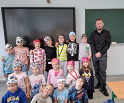 Na zdjęciu widać grupę dzieci wraz z nauczycielem w sali lekcyjnej. Dzieci stoją i siedzą w kilku rzędach, wszystkie mają na głowach kolorowe chusty lub czapki, co nadaje fotografii radosny, wspólnotowy charakter. Uśmiechnięte twarze sugerują, że to pamiątka ze wspólnej zabawy lub wyjątkowego dnia w szkole. W tle znajduje się duży ekran multimedialny i tablica, co potwierdza, że zdjęcie wykonano w klasie.