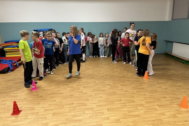 Na zdjęciu widać grupę dzieci biorących udział w zabawie ruchowej w sali gimnastycznej. Dzieci stoją w dwóch rzędach, gotowe do rozpoczęcia konkurencji, a przed nimi ustawione są kolorowe pachołki. W środku sali znajduje się osoba dorosła, prawdopodobnie animatorka lub prowadząca zajęcia, która koordynuje zabawę. W tle widać kolorowe materace oraz ścianę w odcieniach zieleni i bieli. Atmosfera na zdjęciu jest radosna i pełna energii.