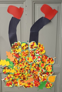 Na zdjęciu widać jesienną dekorację drzwi przygotowaną przez uczniów. Przedstawia ona nogi w czarnych spodniach i czerwonych butach, wystające z dużego, kolorowego stosu jesiennych liści. Liście zostały wykonane z papieru w odcieniach żółci, pomarańczu, czerwieni, brązu i zieleni, co tworzy barwną i radosną kompozycję. Dekoracja jest pomysłowa i pełna jesiennego klimatu – wygląda, jakby ktoś z radością wskoczył w stertę liści.