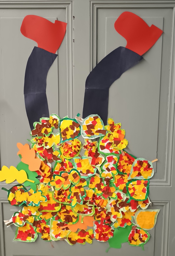 Na zdjęciu widać jesienną dekorację drzwi przygotowaną przez uczniów. Przedstawia ona nogi w czarnych spodniach i czerwonych butach, wystające z dużego, kolorowego stosu jesiennych liści. Liście zostały wykonane z papieru w odcieniach żółci, pomarańczu, czerwieni, brązu i zieleni, co tworzy barwną i radosną kompozycję. Dekoracja jest pomysłowa i pełna jesiennego klimatu – wygląda, jakby ktoś z radością wskoczył w stertę liści.