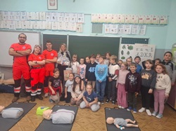 Na zdjęciu widać grupę dzieci oraz dorosłych w sali lekcyjnej. Na pierwszym planie leżą manekiny do nauki pierwszej pomocy, w tym także manekin niemowlęcia, co sugeruje, że odbywają się zajęcia z resuscytacji krążeniowo-oddechowej (RKO).  Z lewej strony stoją trzy osoby w czerwonych mundurach ratowniczych — prawdopodobnie ratownicy medyczni prowadzący zajęcia. Obok nich znajduje się nauczycielka trzymająca bukiet i podziękowania, a przed nimi zgromadzona jest grupa uśmiechniętych dzieci w wieku wczesnoszkolnym.