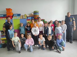 Na zdjęciu widać grupę dzieci oraz dorosłych stojących wspólnie w sali szkolnej. Dzieci ustawione są w kilku rzędach, a po bokach stoją dwaj umundurowani funkcjonariusze Straży Miejskiej. W tle znajdują się kolorowe szafki, pudełka i materiały dydaktyczne. Zdjęcie prawdopodobnie zostało wykonane po zakończeniu spotkania profilaktycznego dotyczącego agresji i przemocy rówieśniczej.