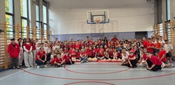 Na zdjęciu widać dużą grupę uczniów i nauczycieli zgromadzonych w sali gimnastycznej. W tle znajduje się kosz do koszykówki oraz ściana z drabinkami gimnastycznymi. Większość osób ma na sobie czerwone koszulki, co sugeruje udział w akcji lub wydarzeniu szkolnym o wspólnym charakterze — np. pokazie pierwszej pomocy, patriotycznym dniu lub inicjatywie integracyjnej.