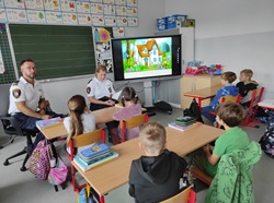 Na zdjęciu widać zajęcia profilaktyczne prowadzone przez funkcjonariuszy Straży Miejskiej w Gliwicach 👮‍♀️👮‍♂️. Dwoje strażników siedzi z przodu klasy i rozmawia z uczniami, którzy z zainteresowaniem słuchają i obserwują prezentację wyświetloną na dużym ekranie. Na monitorze widać kolorową ilustrację z bajki „Siedem koźlątek” 🏡🐐, która stanowi punkt wyjścia do rozmowy o bezpieczeństwie. Dzieci siedzą w ławkach, mają przed sobą zeszyty i plecaki, a w klasie panuje przyjazna, skupiona atmosfera 🌟. Całość wygląda na ciekawą i angażującą lekcję o bezpiecznych zachowaniach.