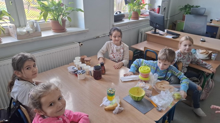 Na zdjęciu widać grupę dzieci siedzących przy stoliku w sali lekcyjnej. Na stole znajdują się różne produkty spożywcze i pojemniki – uczniowie przygotowują swoje zdrowe desery w ramach konkursu „MasterChef Junior”. W tle widać parapety z roślinami doniczkowymi, a cała scena pokazuje miłą i zaangażowaną atmosferę wspólnej pracy.