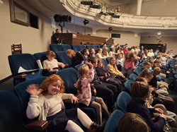 Na zdjęciu widać grupę dzieci siedzących w sali teatralnej. Uczniowie zajmują rzędy miękkich, niebieskich foteli i czekają na rozpoczęcie przedstawienia. Panuje radosna atmosfera — niektóre dzieci rozmawiają ze sobą, inne machają do aparatu lub uśmiechają się. W tle widać balkon oraz stanowisko techniczne z oświetleniem i osobami obsługującymi sprzęt. Scena znajduje się poza kadrem. Zdjęcie oddaje pełne entuzjazmu oczekiwanie młodych widzów na spektakl.