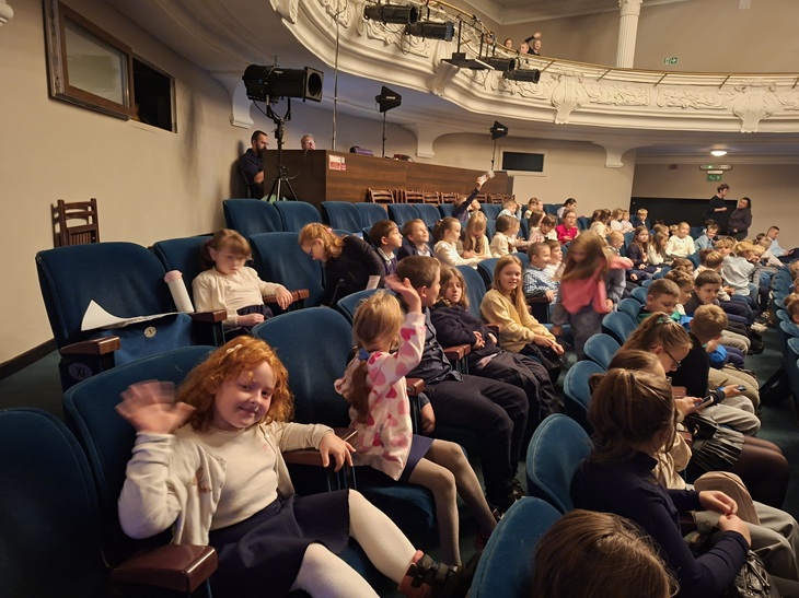 Na zdjęciu widać grupę dzieci siedzących w sali teatralnej. Uczniowie zajmują rzędy miękkich, niebieskich foteli i czekają na rozpoczęcie przedstawienia. Panuje radosna atmosfera — niektóre dzieci rozmawiają ze sobą, inne machają do aparatu lub uśmiechają się. W tle widać balkon oraz stanowisko techniczne z oświetleniem i osobami obsługującymi sprzęt. Scena znajduje się poza kadrem. Zdjęcie oddaje pełne entuzjazmu oczekiwanie młodych widzów na spektakl.