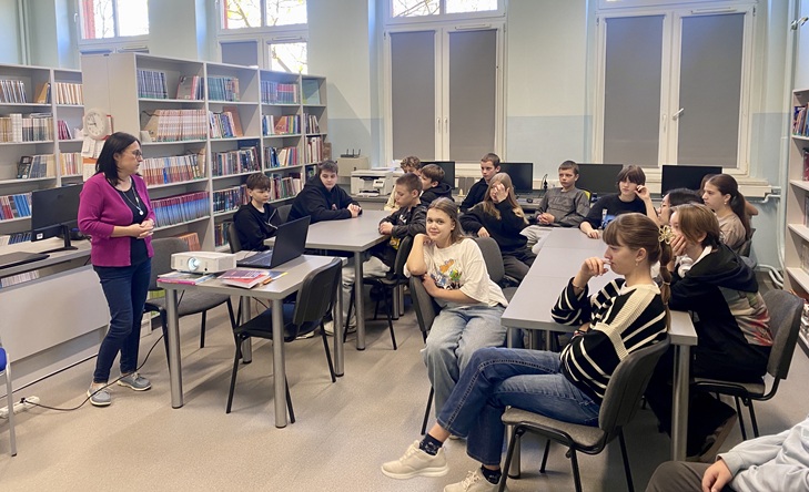 Na zdjęciu widać grupę uczniów siedzących w sali, prawdopodobnie w szkolnej bibliotece lub pracowni informatycznej. Przy stołach siedzi kilkanaście młodych osób, które słuchają kobiety stojącej po lewej stronie — wygląda na nauczycielkę lub prowadzącą zajęcia. Kobieta ma na sobie różowy sweter, ciemne spodnie i białe buty sportowe. Na stole przed nią stoi projektor oraz kilka książek lub notatek. W tle widać regały z książkami oraz kilka komputerów. Sala jest jasna, z dużymi oknami, przez które wpada naturalne światło. Atmosfera wydaje się spokojna i skupiona — uczniowie słuchają z zainteresowaniem.