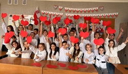 Na zdjęciu widać grupę dzieci w szkolnej sali, które wspólnie świętują 11 listopada – Święto Niepodległości. Uczniowie ubrani są głównie na biało, co nawiązuje do barw narodowych Polski. W dłoniach trzymają czerwone papierowe serca, które unoszą w górę, tworząc radosną i patriotyczną atmosferę. W tle na ścianie widać ozdoby i napisy w biało-czerwonych barwach, m.in. girlandę z napisem „11 LISTOPADA ŚWIĘTO NIEPODLEGŁOŚCI”. Cała scena jest pełna energii i uśmiechów, oddając radosny charakter szkolnych obchodów tego narodowego święta.