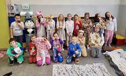 Na zdjęciu widać grupę dzieci pozujących w sali szkolnej podczas tematycznego dnia, prawdopodobnie Dnia Piżamy. Wszystkie dzieci ubrane są w kolorowe piżamy lub kombinezony z motywami zwierząt i bajkowych postaci. Niektóre trzymają pluszowe maskotki, a kilka osób robi zabawne pozy, jakby spały lub przytulały pluszaki.