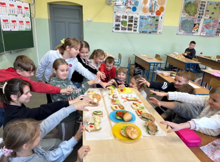 Na zdjęciu widać grupę dzieci siedzących wokół długiego stołu w szkolnej klasie. Na środku stołu leżą talerze i tacki z kolorowymi, zdrowymi kanapkami oraz warzywami. Dzieci z uśmiechami wyciągają ręce w stronę przygotowanych potraw, sprawiając wrażenie, jakby wspólnie celebrują chwilę przed rozpoczęciem posiłku.