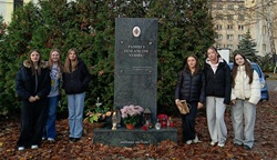 Na zdjęciu widoczna jest grupa sześciu młodych osób stojących po obu stronach pomnika Pamięci Zesłańców Sybiru. Pomnik jest wykonany z ciemnego kamienia, ustawiony na tle gęstych, zielonych krzewów. U jego podstawy znajdują się zapalone znicze, kwiaty oraz drobne pamiątkowe dekoracje, co tworzy atmosferę zadumy i szacunku.