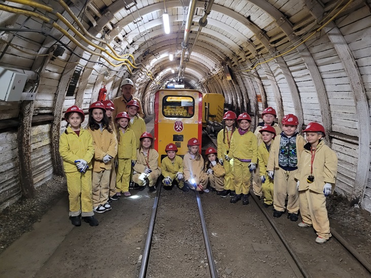 Na zdjęciu widać grupę dzieci ubranych w żółte kombinezony ochronne i czerwone kaski górnicze. Stoją one w tunelu kopalnianym, obok małej górniczej kolejki o żółto-czerwonych barwach. W tle widać oświetlony korytarz z metalowymi obudowami i żółtymi przewodami biegnącymi wzdłuż ścian.