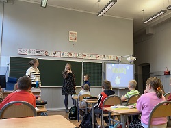 Na zdjęciu widać zajęcia prowadzone w klasie szkolnej. Przy tablicy stoją dwie osoby dorosłe – prawdopodobnie pedagog i psycholog – które rozmawiają z uczniami. Jedno z dzieci uczestniczy aktywnie w zajęciach, stojąc przy prowadzących, podczas gdy pozostali uczniowie siedzą w ławkach i uważnie słuchają.