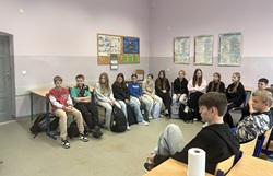 Na zdjęciu widać grupę uczniów siedzących w kręgu w sali lekcyjnej. Młodzież uważnie słucha prowadzącego, co sugeruje, że uczestniczą w warsztatach lub zajęciach dyskusyjnych. W tle znajdują się tablice ścienne z plakatami edukacyjnymi oraz tablica korkowa z informacjami szkolnymi. Uczniowie wyglądają na zaangażowanych i skupionych, a atmosfera w sali wydaje się spokojna i przyjazna. Zdjęcie dobrze oddaje charakter zajęć poświęconych budowaniu relacji i współpracy w grupie.