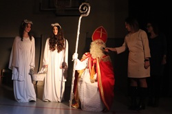 Na zdjęciu widoczna jest scena świątecznego występu. W centrum znajduje się Święty Mikołaj w tradycyjnym stroju, trzymający pastorał i przemawiający do publiczności. Obok stoją dwie osoby przebrane za anioły w białych szatach, a z prawej strony osoba z mikrofonem. Całość odbywa się na scenie z przyciemnionym tłem, co podkreśla uroczysty i świąteczny charakter wydarzenia.