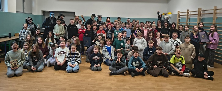 Na zdjęciu widać dużą, liczną grupę uczniów pozujących wspólnie w sali gimnastycznej. Dzieci siedzą i stoją w kilku rzędach, uśmiechają się i prezentują energiczną, radosną atmosferę po zakończonej grze miejskiej.  W tle stoją także przebrani aktorzy – ich stroje są wyraziste i teatralne, co dodaje zdjęciu klimatu zabawy i przygody. Uczniowie mają na sobie codzienne, kolorowe ubrania, a część z nich trzyma w rękach upominki otrzymane po wydarzeniu. Cała scena wygląda na pełną entuzjazmu i pozytywnych emocji, jak po dobrze spędzonym, pełnym atrakcji dniu.