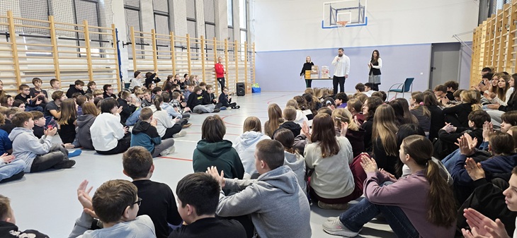 Na zdjęciu widać uczniów zgromadzonych w sali gimnastycznej, siedzących na podłodze i słuchających prowadzących stojących na środku. Atmosfera wygląda na uroczystą, a uczniowie biją brawo.