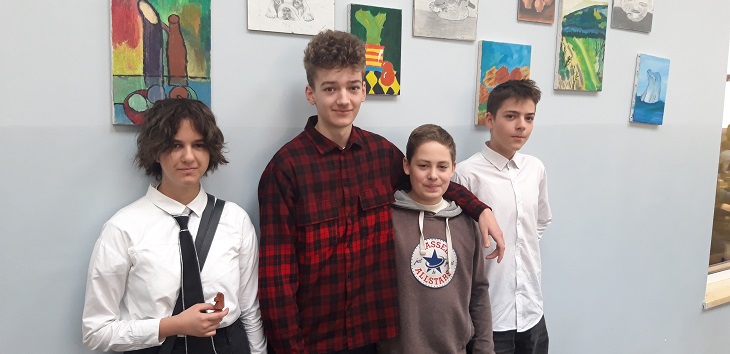 Na zdjęciu widać czworo młodych uczniów stojących obok siebie na szkolnym korytarzu. W tle wiszą kolorowe prace plastyczne.  Osoba po lewej ma krótkie, kręcone włosy, jest ubrana w białą koszulę i czarny krawat.  Obok stoi wysoka osoba w czerwono-czarnej koszuli w kratę, z lekkim uśmiechem.  Trzecia osoba ma na sobie jasną bluzę z kapturem i stoi bardzo blisko poprzedniej osoby, obejmując ją ramieniem.  Po prawej stronie stoi czwarta osoba w białej koszuli, patrząca lekko w bok.