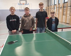 Na zdjęciu widać czterech chłopców stojących przy stole do tenisa stołowego. Wszystkie pozują do zdjęcia, uśmiechając się delikatnie lub patrząc spokojnie w stronę aparatu. Z przodu znajduje się stół pingpongowy z siatką, piłeczką oraz dwoma rakietkami leżącymi po obu stronach stołu.