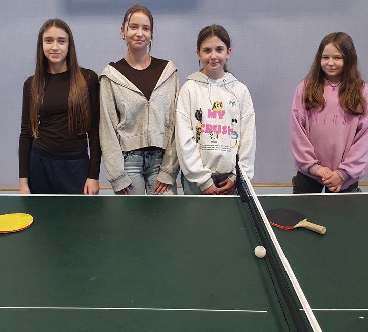 Na zdjęciu widać cztery dziewczyny stojące przy stole do tenisa stołowego. Wszystkie pozują do zdjęcia, uśmiechając się delikatnie lub patrząc spokojnie w stronę aparatu.  Z przodu znajduje się stół pingpongowy z siatką, piłeczką oraz dwoma rakietkami leżącymi po obu stronach stołu.