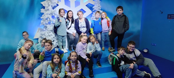Na zdjęciu widać grupę dzieci wraz z opiekunkami, pozujących do wspólnej fotografii w Funzeum. Tło zdjęcia jest niebieskie, ozdobione białymi, sztucznymi drzewkami i dużym symbolem śnieżynki, co tworzy zimową, bajkową scenerię